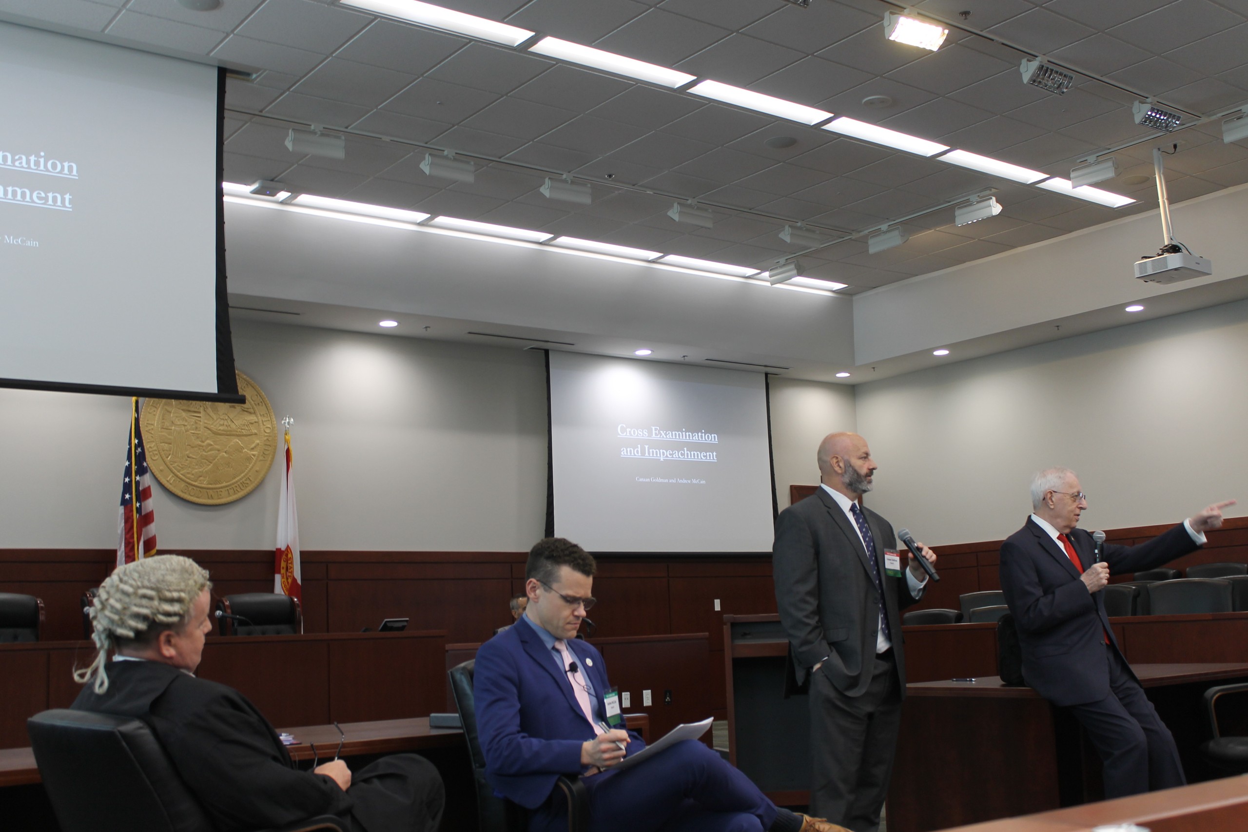 A group of individuals is engaged in a discussion in a formal setting, with one person speaking behind a lectern while others listen attentively. A presentation display is visible in the background. Flags and a seal are present in the room.