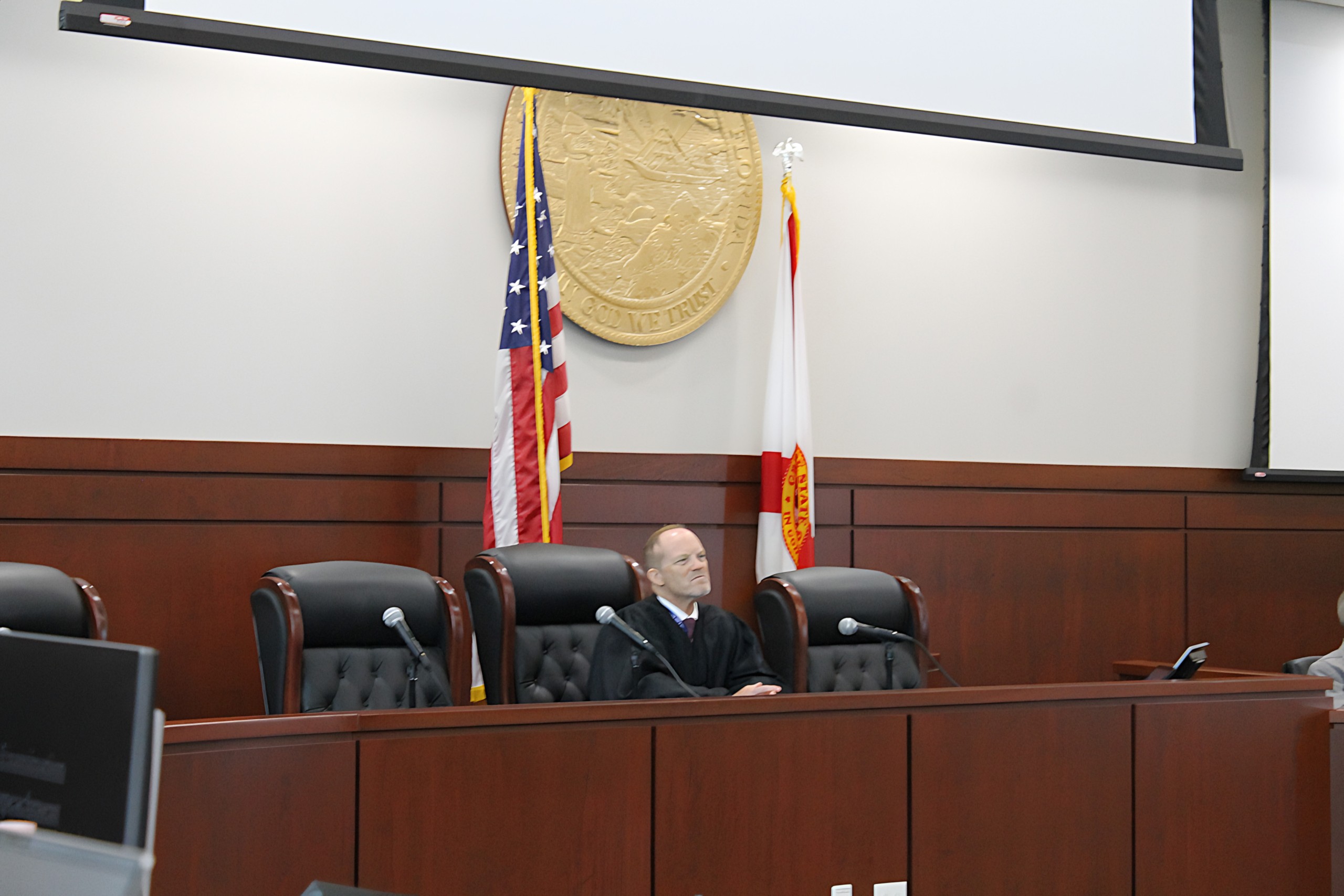 An individual seated behind a bench in a courtroom, with flags and a seal displayed in the background.