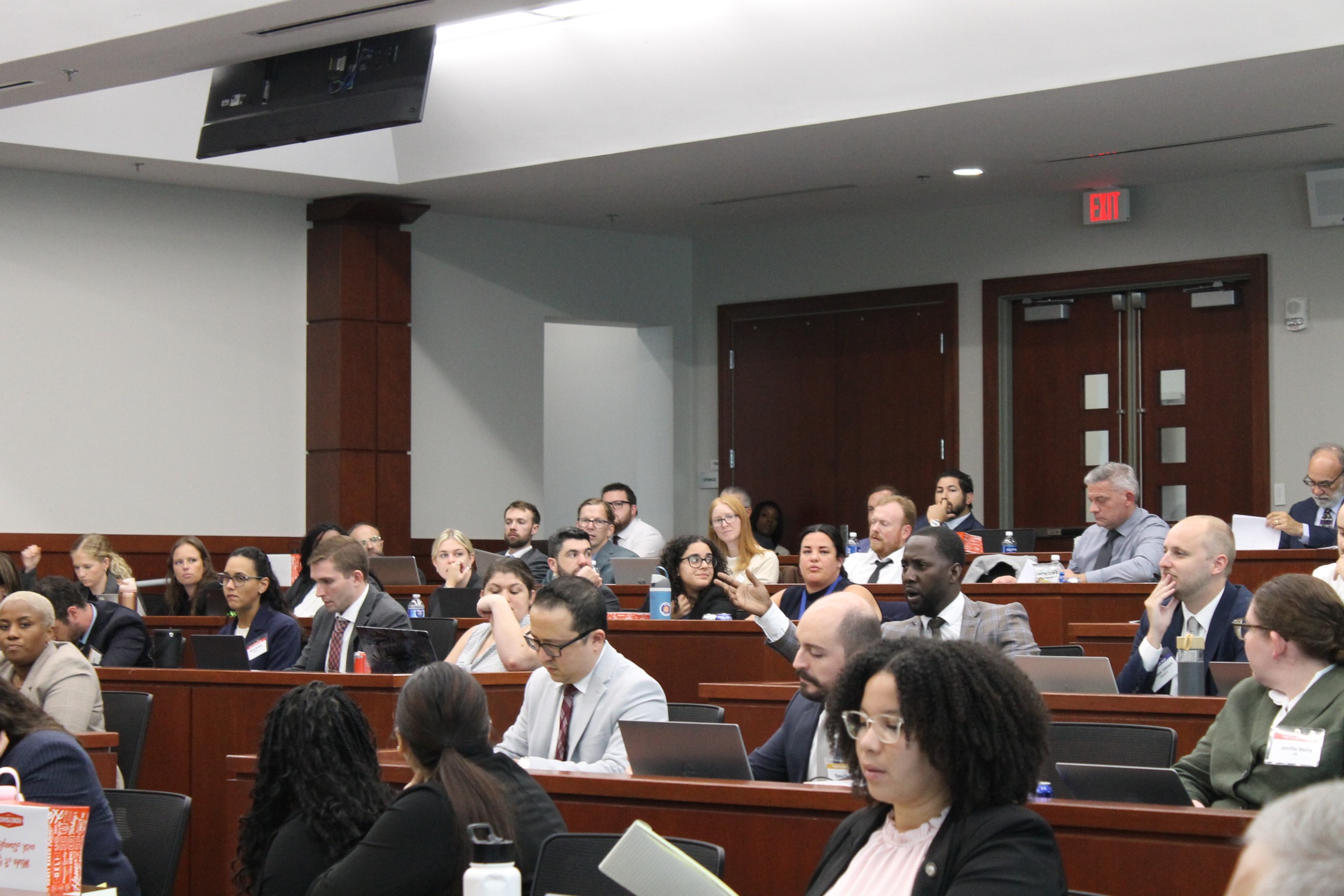 A speaker presents to an audience seated in a courtroom. Some attendees are engaged with laptops or taking notes. The room features wooden paneling and multiple doors in the background.