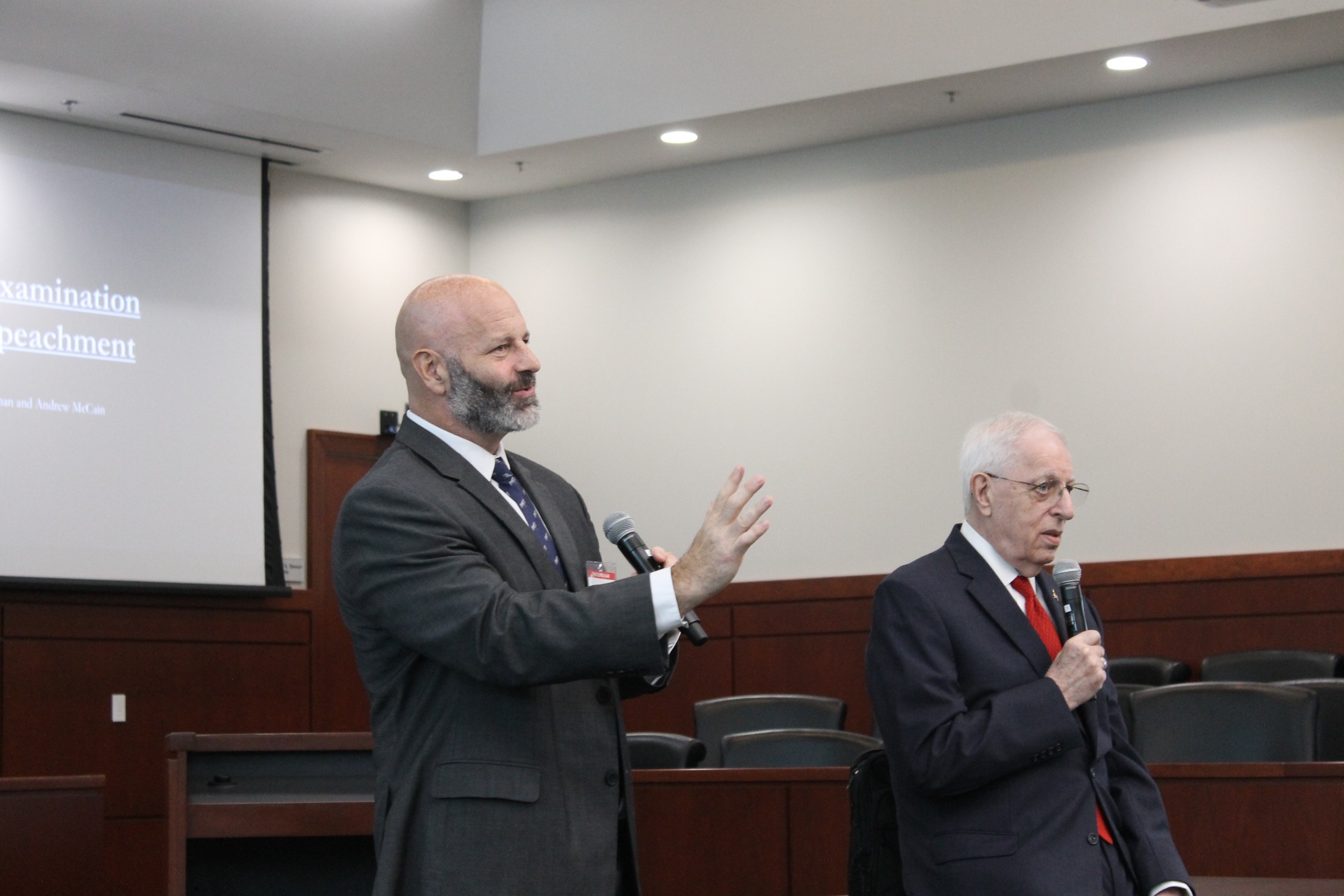 Two speakers are addressing an audience. One speaker gestures while holding a microphone, and the other speaker listens attentively. A presentation screen is visible in the background.