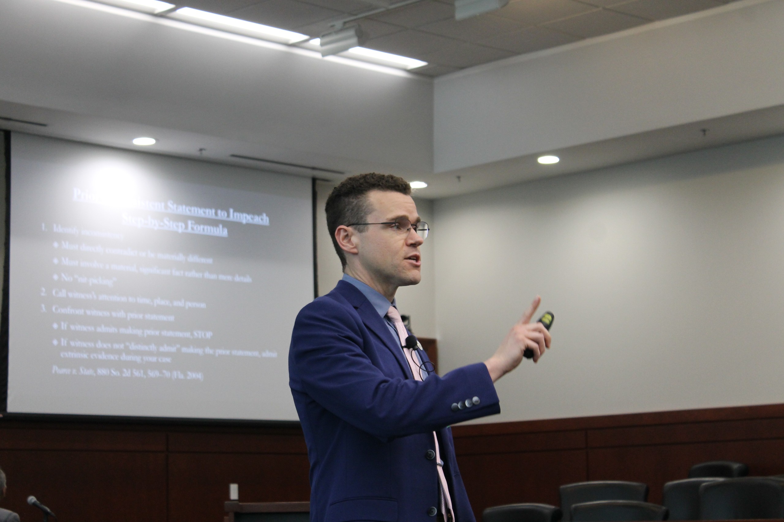 A person is speaking in front of an audience. A presentation is visible on the screen behind, containing bullet points related to a step-by-step formula for making a statement to impeach.

