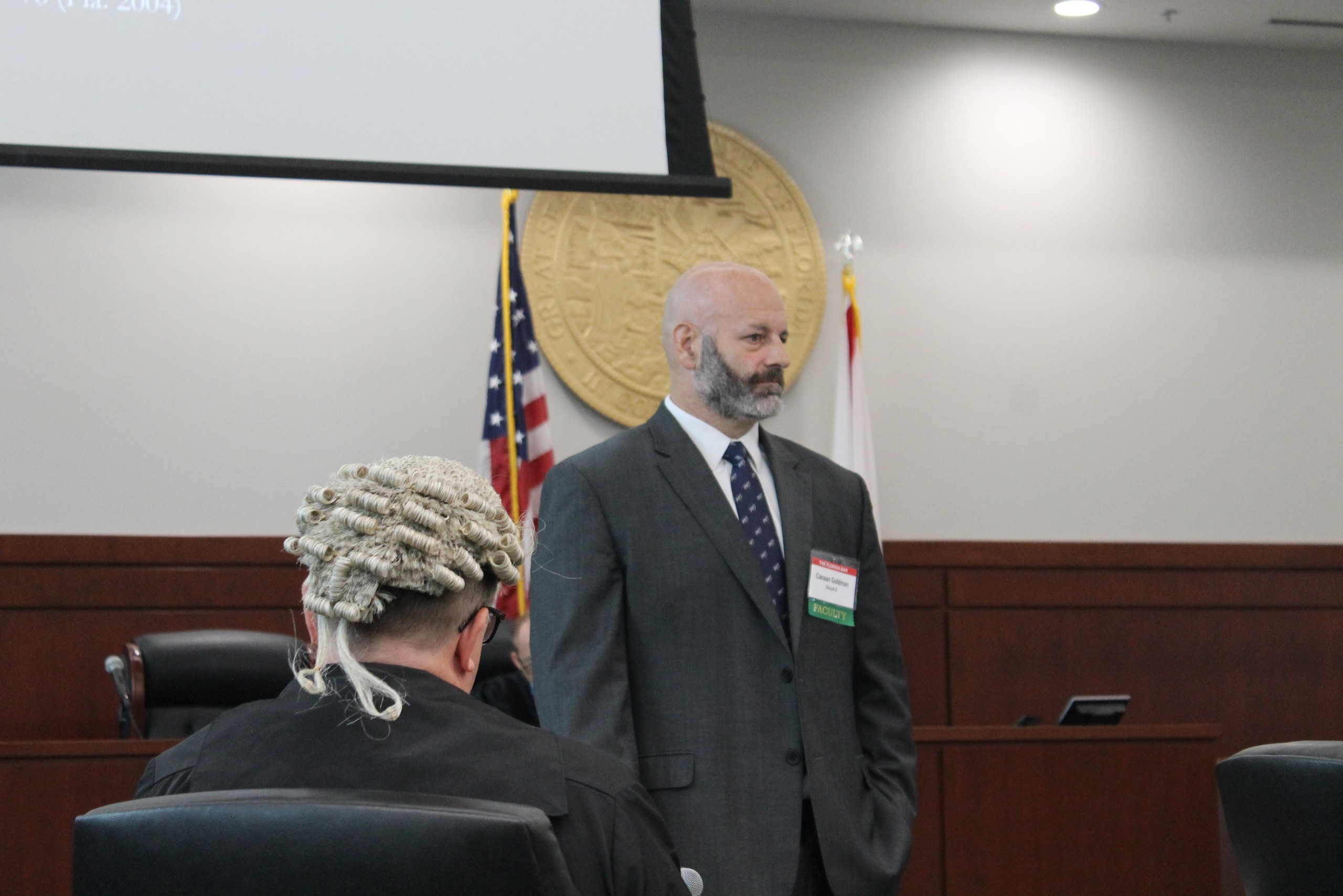 A person in formal attire speaks in a courtroom setting, with flags and a seal visible in the background. Another individual, seated and wearing court attire, is partially visible in the foreground.