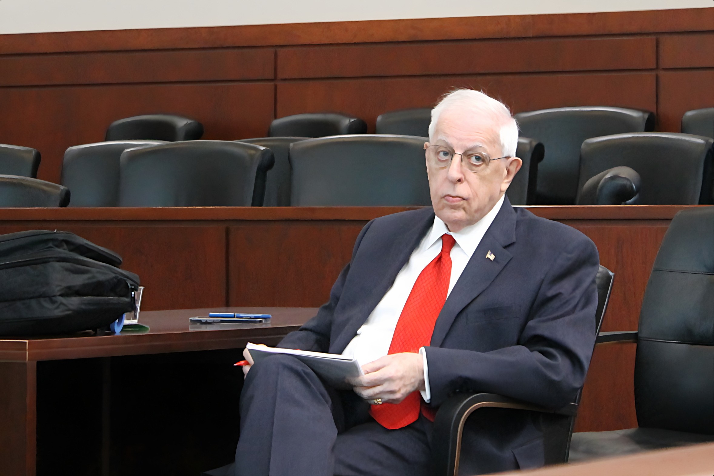 A person seated at a table in a courtroom, holding a notebook and looking towards the camera.