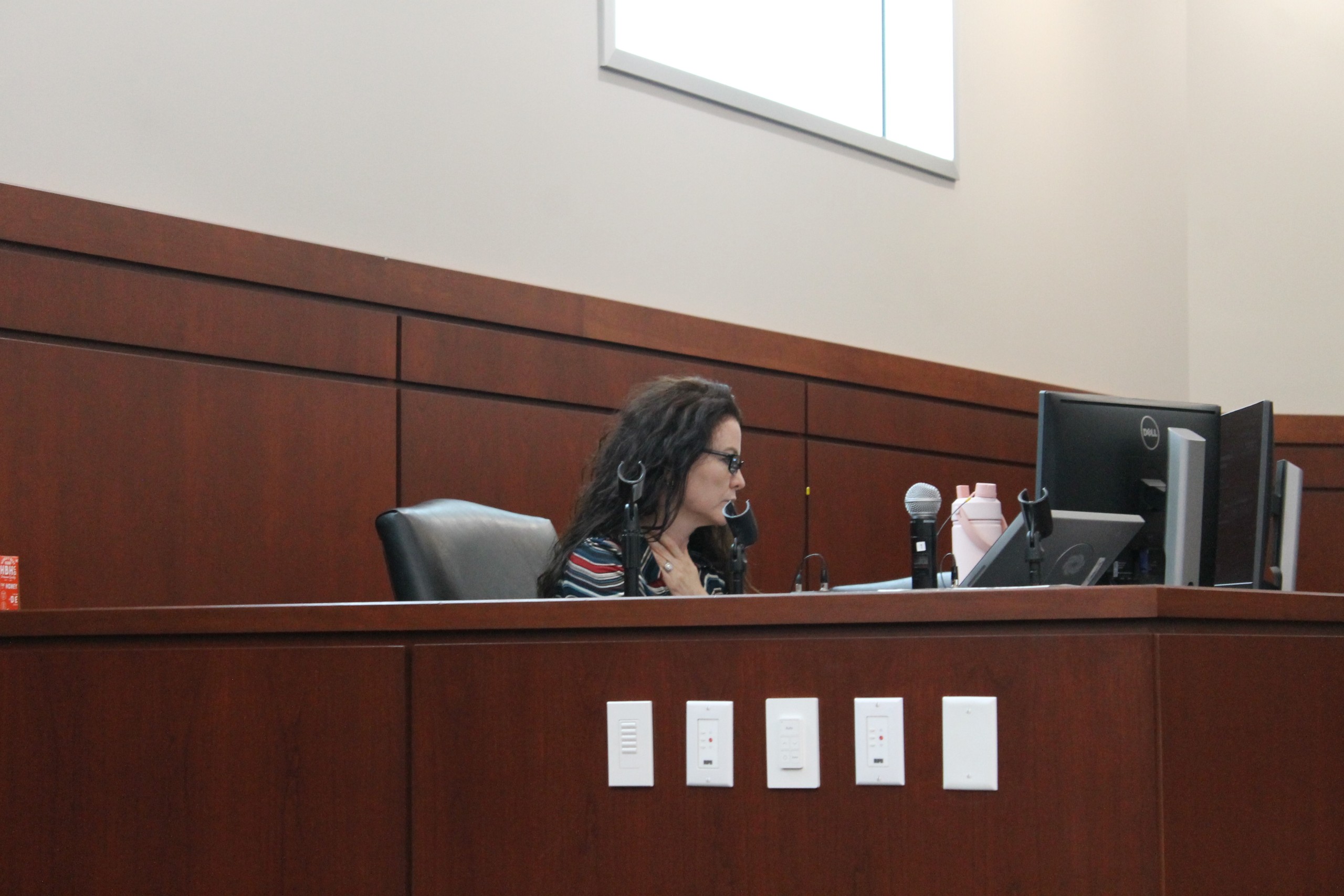 A person sits at a desk in a formal setting, focused on a computer, with a microphone and a water bottle nearby.
