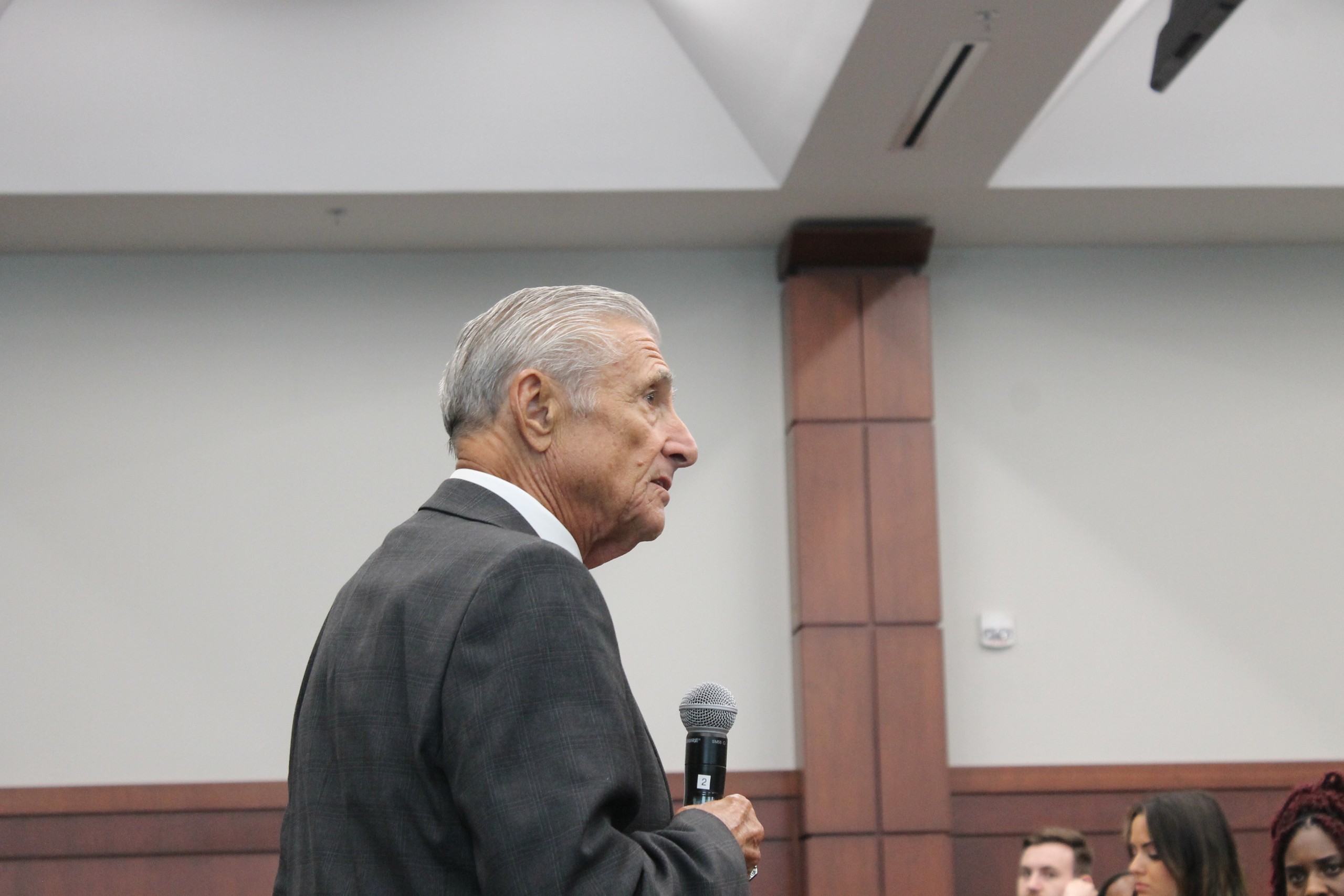 An individual speaks into a microphone while standing in a room filled with people. The speaker faces to the side, with a wood-paneled wall in the background.