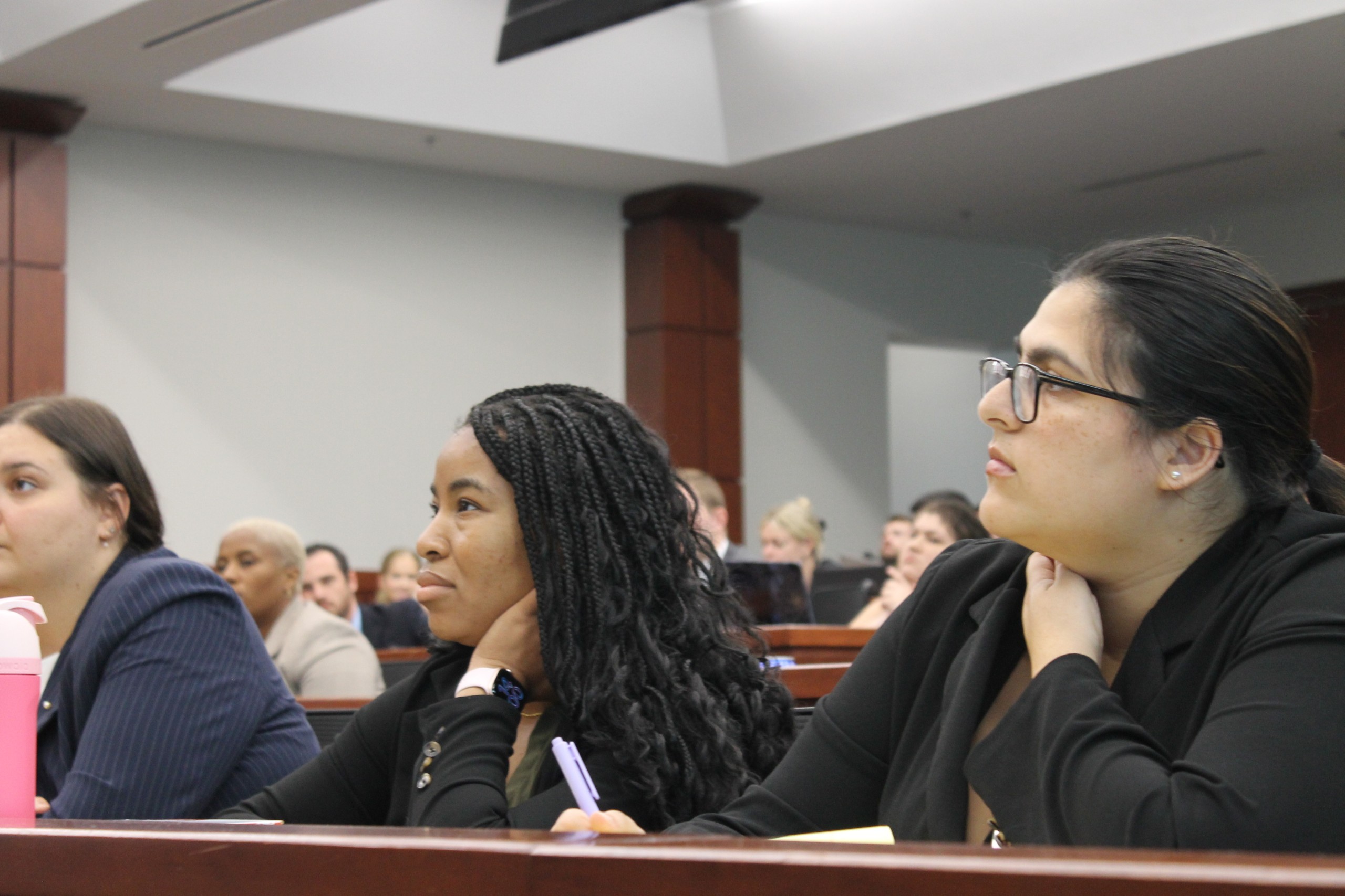 A group of individuals seated and attentively listening in a classroom setting. A few people are visible in the foreground, with others in the background engaged in the session. The environment appears formal, likely part of a lecture or discussion.
