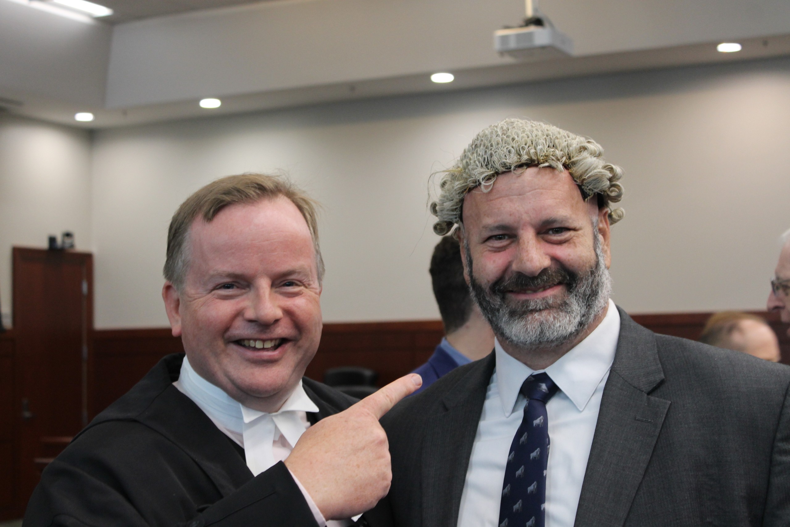 Two individuals pose together, smiling in a courtroom setting. One is dressed in a dark robe, while the other wears formal attire with a light-colored tie. The background features wooden paneling and blurred figures of others present.