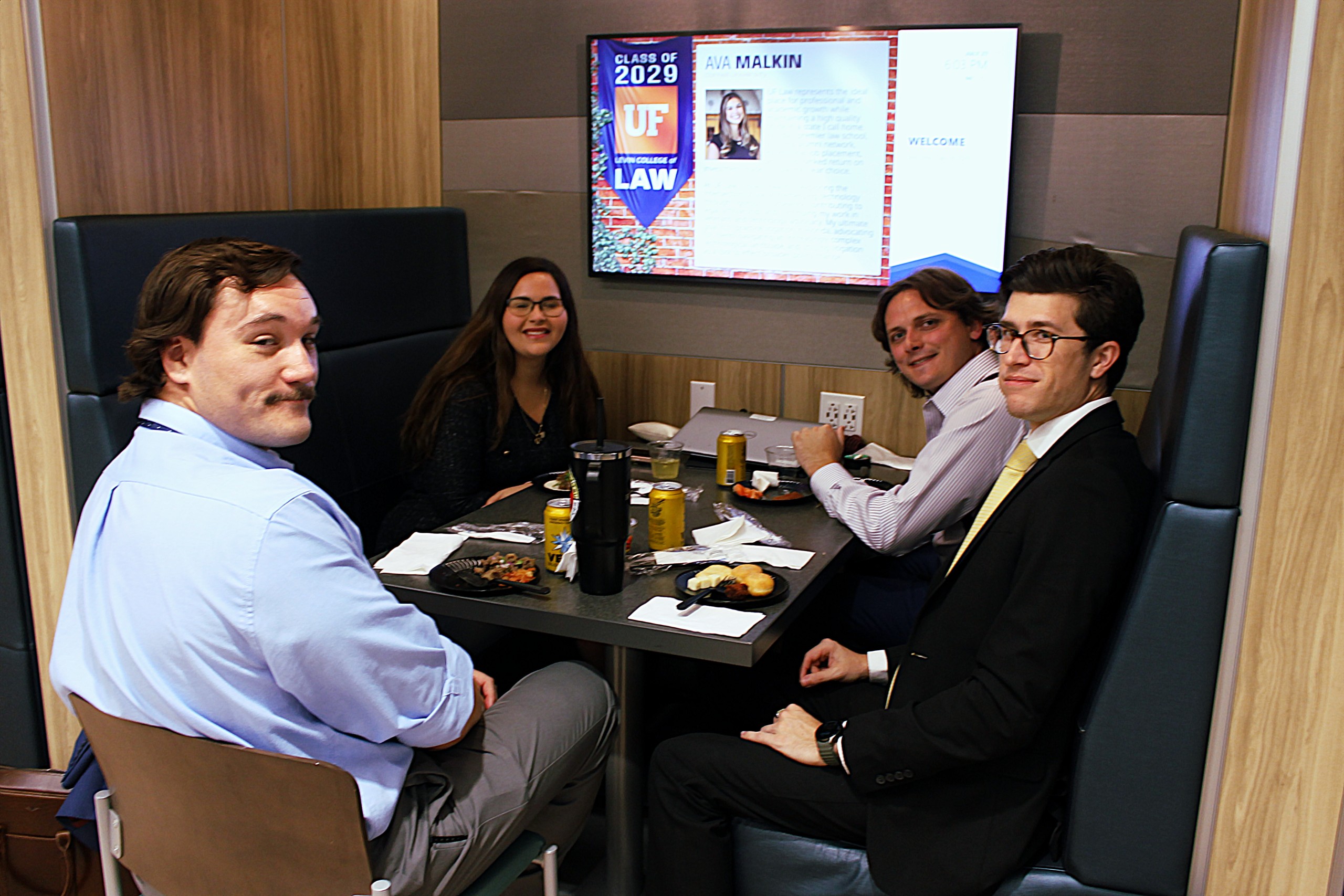 Four individuals are seated at a table with food and drinks. A screen in the background displays information about a law program. The setting appears to be a casual gathering or meeting.