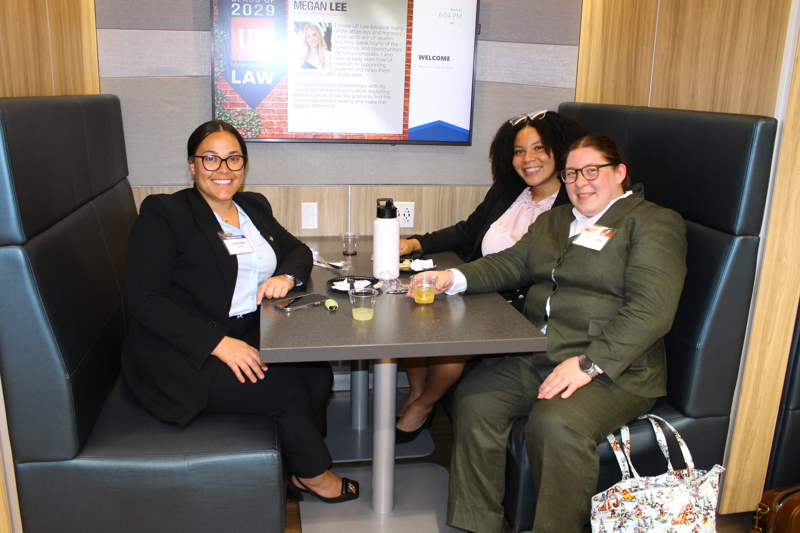Three individuals sit together at a table in a booth, smiling and engaged in conversation. They are dressed in business attire and have drinks in front of them. A screen in the background displays information related to an event.