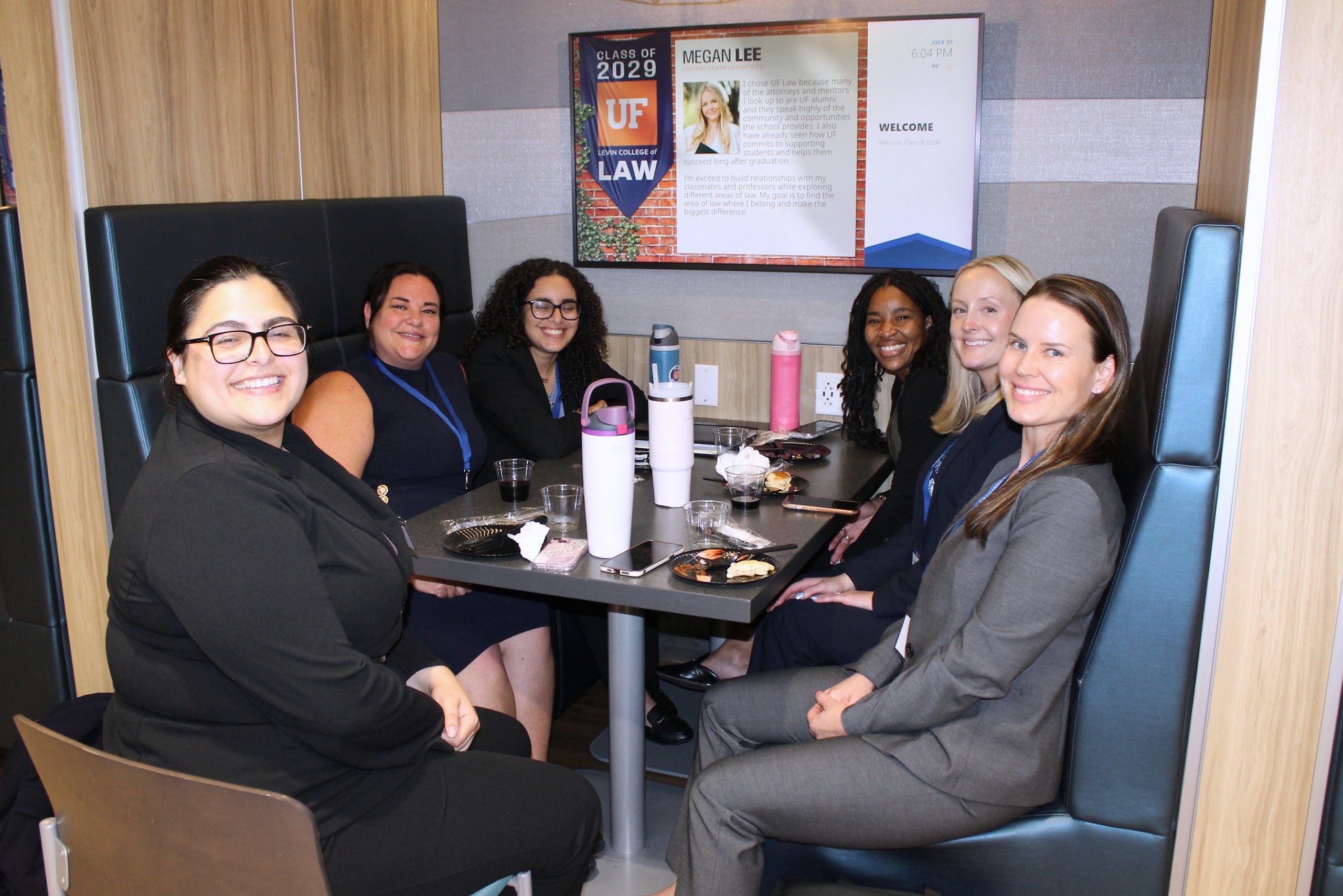 A group of six individuals sits together at a table in a cozy setting, smiling and enjoying food and beverages. A screen on the wall displays information about an event.