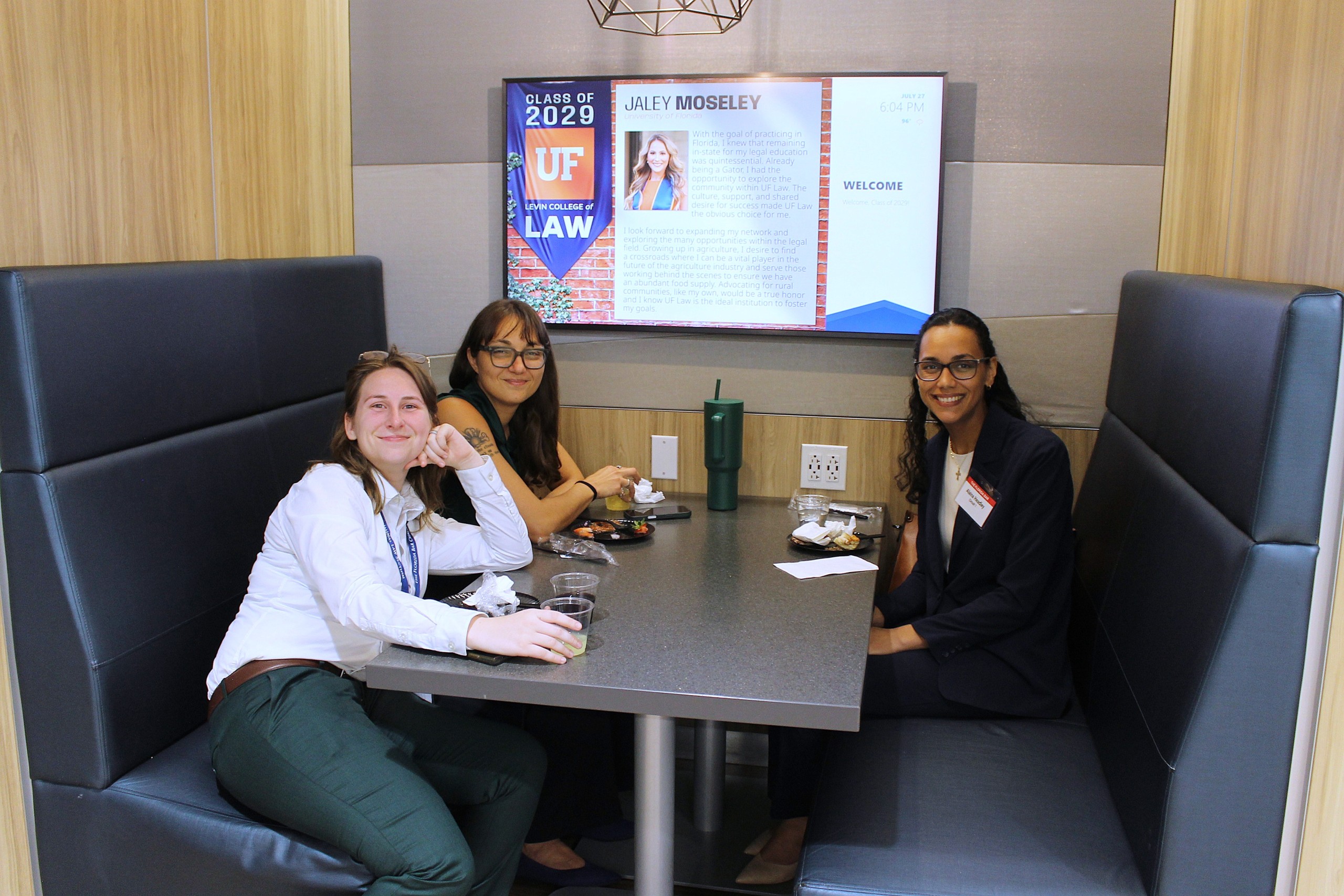 Three individuals are seated at a table in a booth enjoying food and drinks. Behind them is a screen displaying information about UF Law.