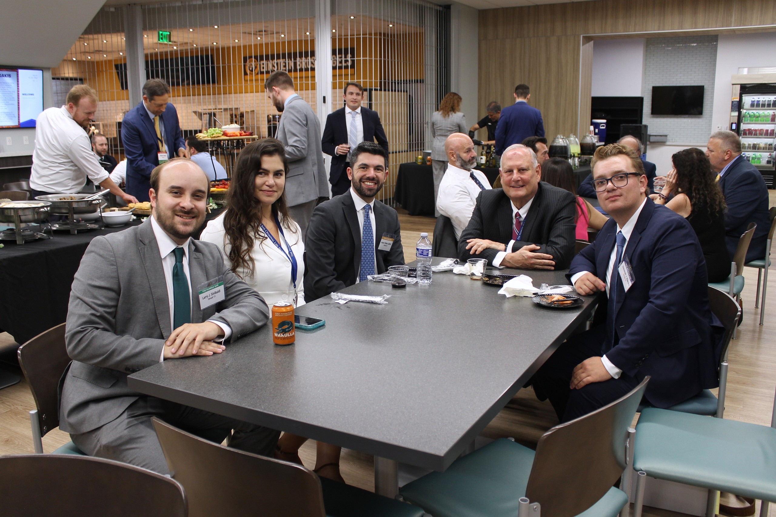 A group of five individuals sit around a table in a dining area, smiling and engaging in conversation. There are various food items on the table, and several people can be seen in the background, mingling and serving food.