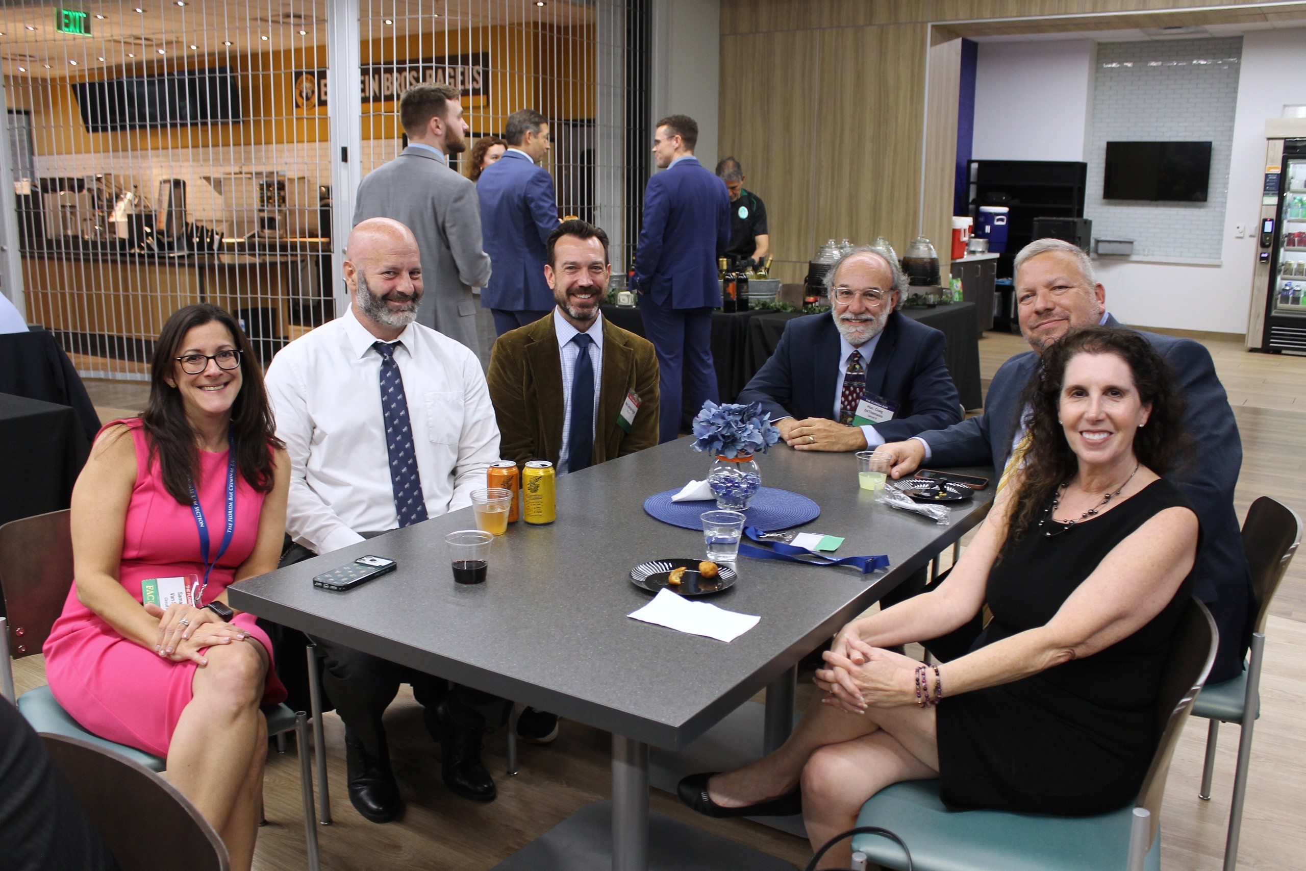 A group of six people sits around a table with snacks and drinks in a well-lit, casual setting. They appear to be engaged in conversation, with one person holding a phone. In the background, additional individuals can be seen mingling.