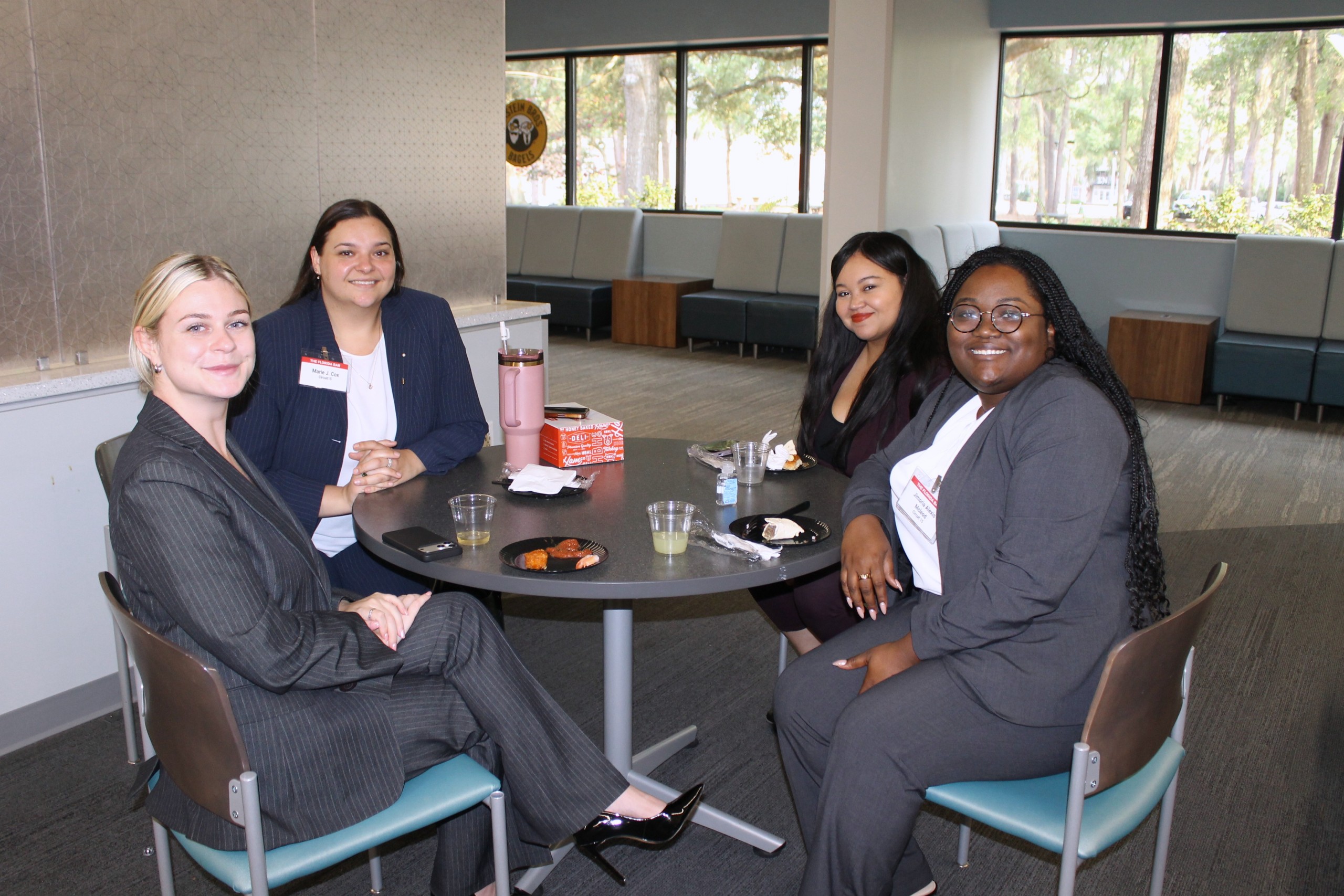 Four individuals sit around a table with refreshments, smiling and engaged in conversation. The setting features large windows and comfortable seating arrangements in the background.