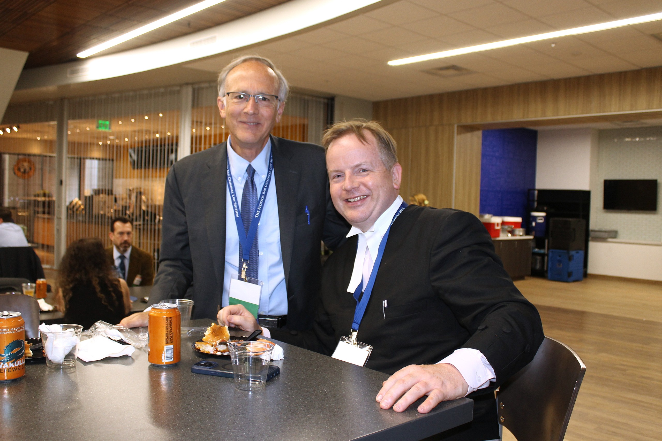 Two individuals pose together at a table in a spacious, modern indoor setting. They are smiling and wearing name tags. In the foreground, there are snacks and drinks on the table. The background shows a casual gathering, with other people and seating visible.