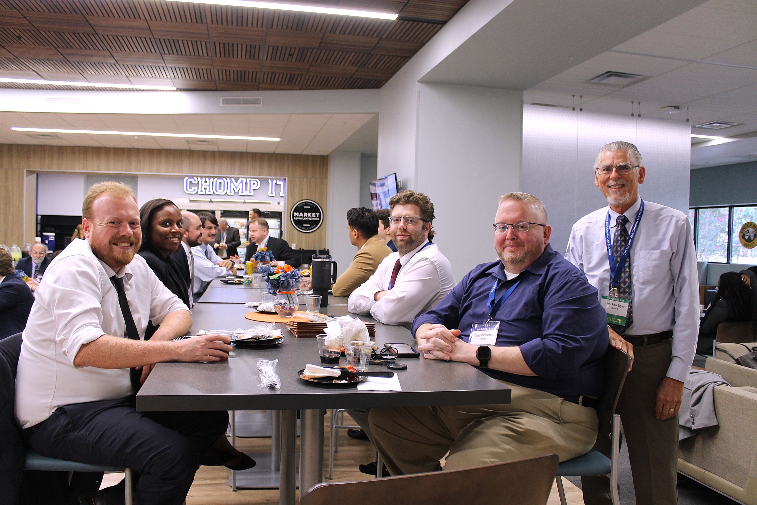 A group of individuals sits at a table in a dining area, with some engaging in conversation. Food and drinks are on the table. In the background, several people can be seen mingling. The setting appears lively and social.