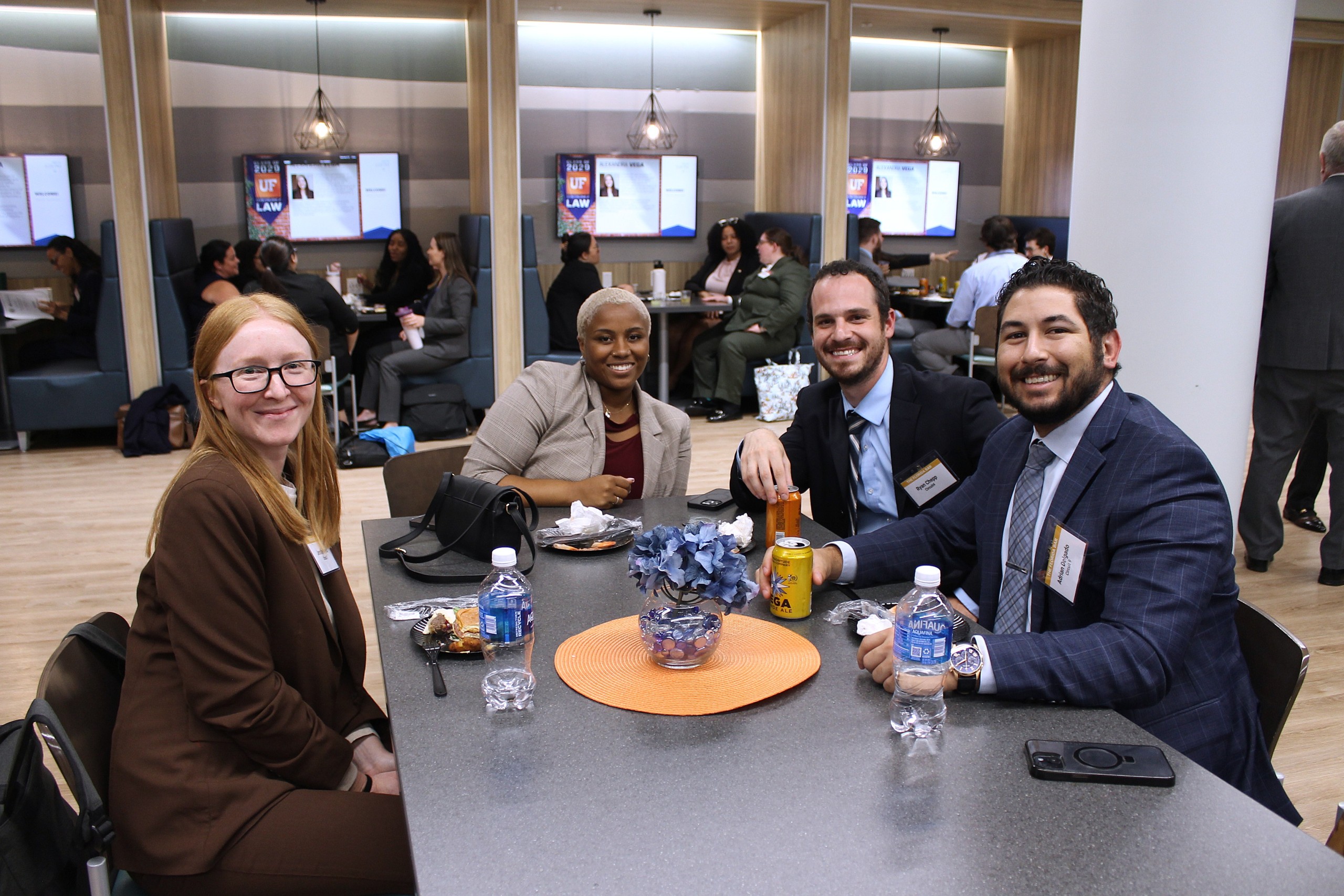 A group of four individuals sits around a table sharing food and drinks in a modern space. In the background, others are engaged in conversation. Screens display information related to an event.