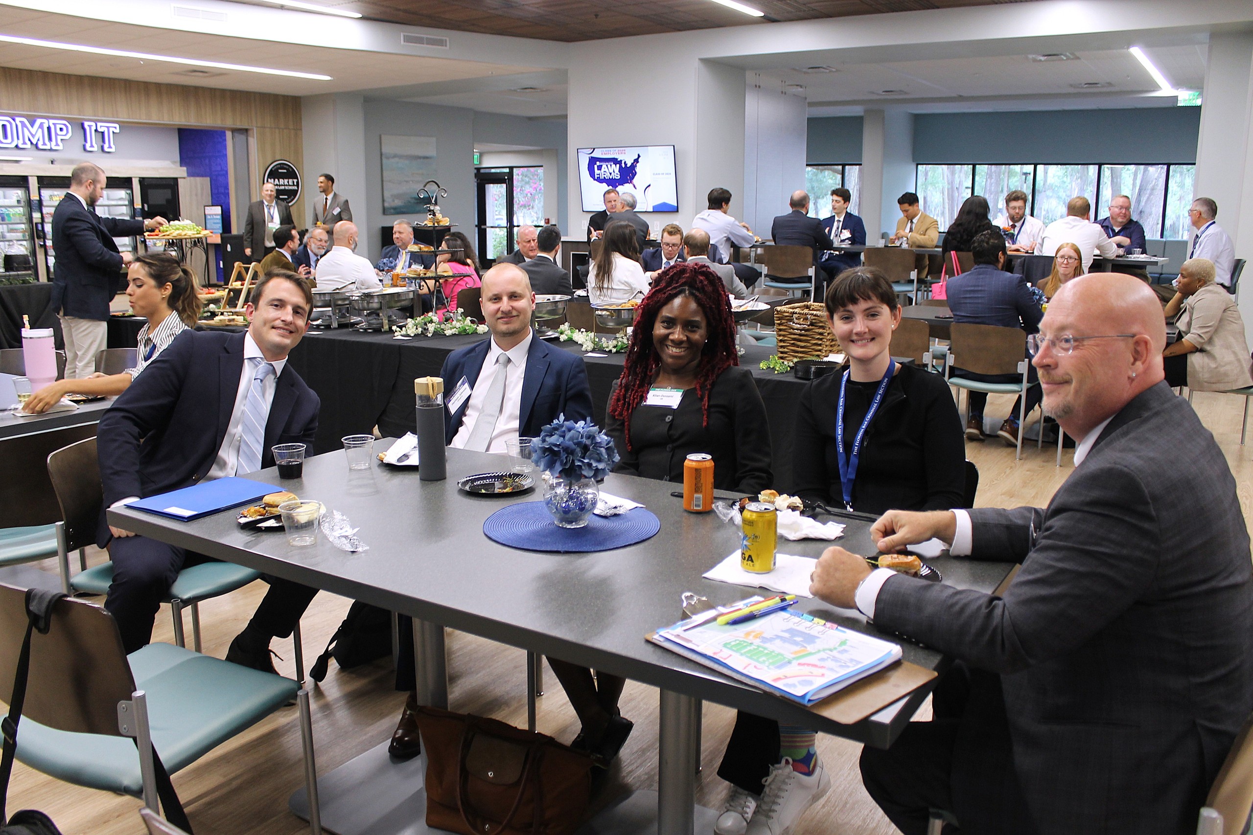 A group of five people is seated at a table in a modern gathering space, engaging in conversation and enjoying refreshments. In the background, additional attendees mingle and network. A food display is visible, along with a digital screen showing a map.