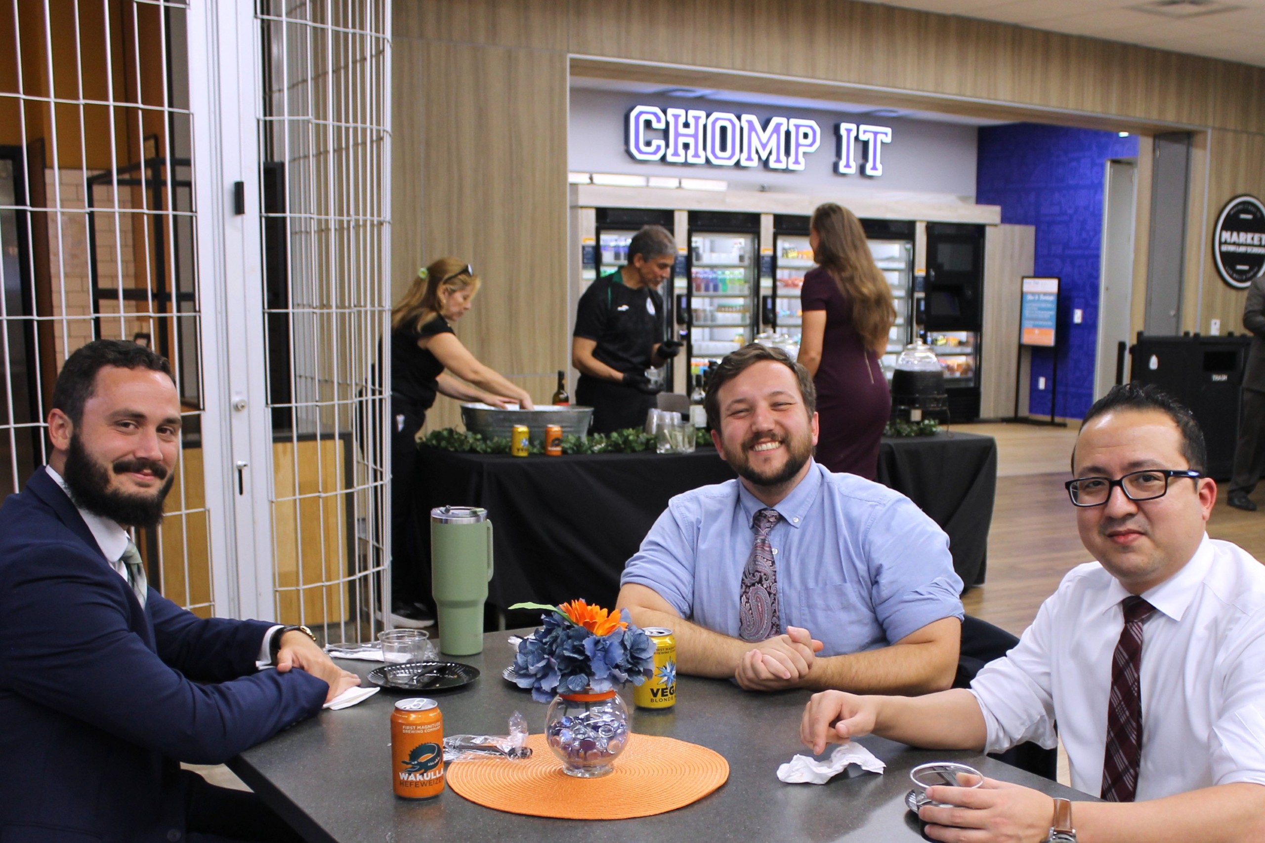 Three individuals sit together at a table, smiling and engaging with each other. The table is decorated with a small flower arrangement and a colorful placemat. In the background, others are preparing food.