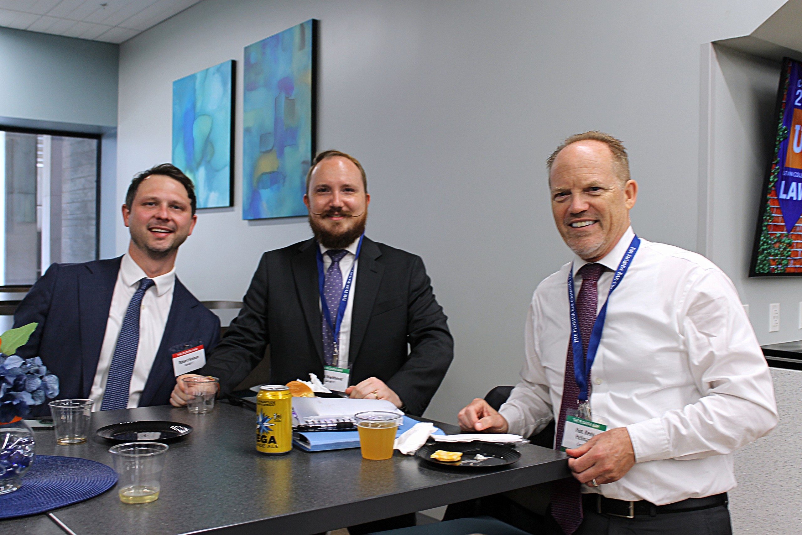 Three individuals are seated at a table in a modern setting, smiling and engaging with each other. They are dressed in formal attire and have drinks and snacks on the table. Colorful artwork is visible on the wall behind them.